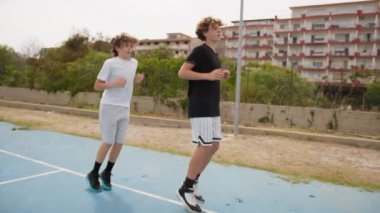İki genç erkek dışarıda mavi alanda basketbol oynuyorlar..