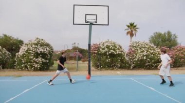 İki genç erkek dışarıda mavi alanda basketbol oynuyorlar..