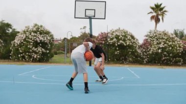 İki genç erkek dışarıda mavi alanda basketbol oynuyorlar..