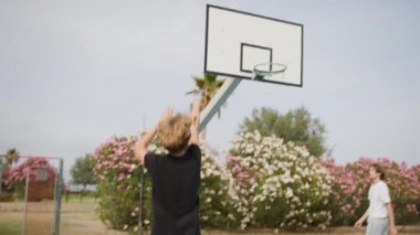 İki genç erkek dışarıda mavi alanda basketbol oynuyorlar..