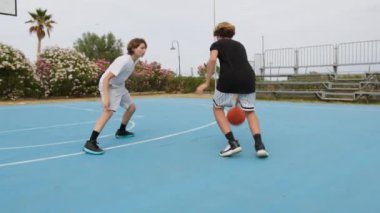 İki genç erkek dışarıda mavi alanda basketbol oynuyorlar..