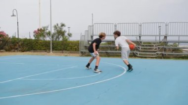 İki genç erkek dışarıda mavi alanda basketbol oynuyorlar..