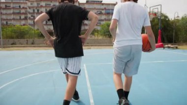 İki genç erkek dışarıda mavi alanda basketbol oynuyorlar..
