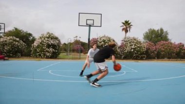 İki genç erkek dışarıda mavi alanda basketbol oynuyorlar..