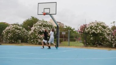 İki genç erkek dışarıda mavi alanda basketbol oynuyorlar..