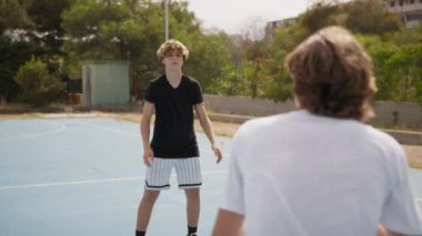İki genç erkek dışarıda mavi alanda basketbol oynuyorlar..