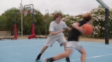 İki genç erkek dışarıda mavi alanda basketbol oynuyorlar..