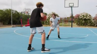 İki genç erkek dışarıda mavi alanda basketbol oynuyorlar..