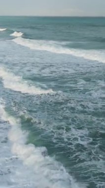 Plaja çarpan sıvı dalgaların hava görüntüsü, rüzgarla karıştırılmış ve okyanus ufku boyunca büyüleyici bir manzara yaratıyor.