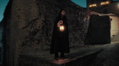 Gizemli genç kadın pelerin ve fenerle bir köyün sokaklarında yürüyor..