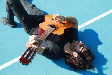 Yerde yatan adam mavi zeminde klasik gitarı çalıyor..