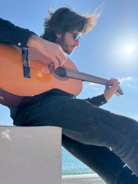 Uzun Saçlı Çocuk Yazın Okyanusta Siyah Giyinik Klasik Gitar Çalıyor.