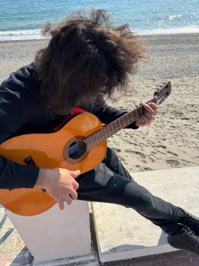 Uzun Saçlı Çocuk Yazın Okyanusta Siyah Giyinik Klasik Gitar Çalıyor.