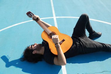 Yerde yatan adam mavi zeminde klasik gitarı çalıyor..