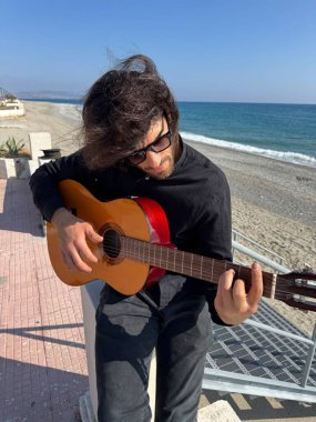 Uzun Saçlı Çocuk Yazın Okyanusta Siyah Giyinik Klasik Gitar Çalıyor.