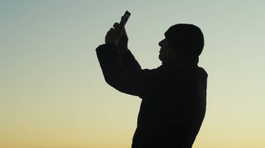 Gün batımında akıllı telefonuyla fotoğraf çeken bir adamın silueti..