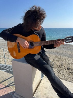 Uzun Saçlı Çocuk Yazın Okyanusta Siyah Giyinik Klasik Gitar Çalıyor.