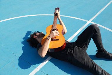 Yerde yatan adam mavi zeminde klasik gitarı çalıyor..