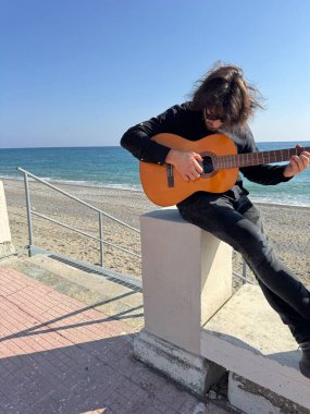 Uzun Saçlı Çocuk Yazın Okyanusta Siyah Giyinik Klasik Gitar Çalıyor.