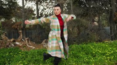 Ekose ceketli kadın, baharda ağaçlı ve sarı çiçekli bir bahçede dans ediyor..