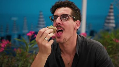 Adam hamburger yiyor ve arka planda deniz kenarında gülümsüyor. . 