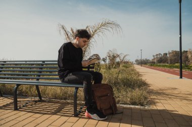 Doğada bir bankta oturan bir adam sırt çantasında bir şey arıyor. . 