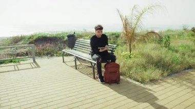 Deniz kenarındaki bankta oturan genç adam sırt çantasından bir defter seçiyor. .