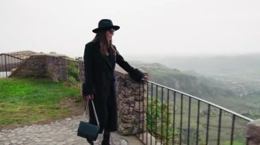 Bir kadın yürüyor ve korkulukların arkasındaki manzarayı izliyor. .