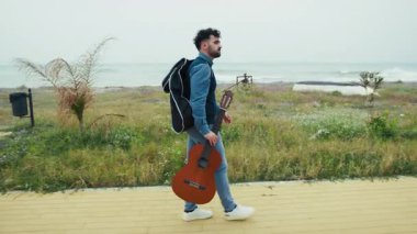 Bir adam elinde gitarıyla denizin kenarındaki kaldırımda yürüyor.. 