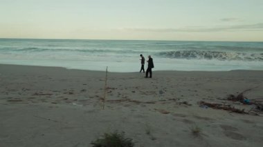 Uzun bir sahilde, iki insan okyanus dalgalarının öfkesini izlerken yürür..