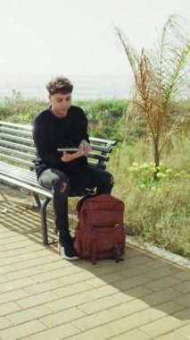 Deniz kenarındaki bankta oturan genç adam sırt çantasından bir defter seçiyor. .