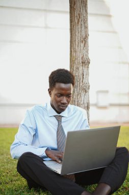 African man working with his computer. He is outdoors sitting on grass next to a tree.