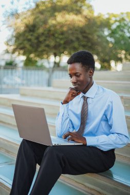 Smiling african businessman working with his laptop. He is sitting on stairs.