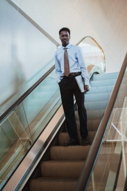 Happy african businessman holding a laptop while is going down on a escalator.