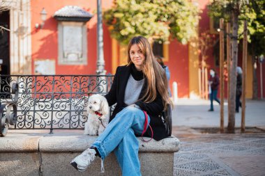 Teenager looking at camera, sitting next to her white dog. They are sitting on a stone bench in the city.