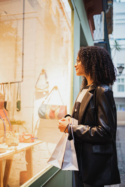 Улыбающаяся девушка смотрит в витрину магазина, в то время как она держит некоторые сумки для покупок