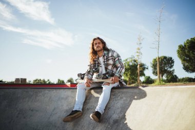 Ciddi bir genç adamın kameraya bakarken çekilmiş fotoğrafı. Bir kaykay parkında oturuyor ve elinde bir kaykay tutuyor.