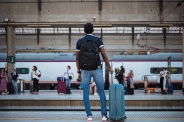 Treni bekleyen Afrikalı bir adamın arka planı. Elinde mavi bir bavul ve siyah bir çanta var..