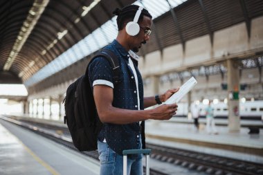Afrikalı bir adamı tren istasyonunda harita okurken çekmişler. O müzik dinlerken bekliyor.