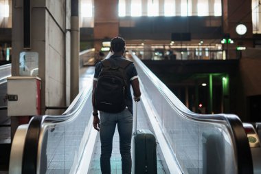 Delikanlının, tren istasyonunda yürüyen merdivenle giderken elinde bavul tutarken çekilmiş bir fotoğrafı.