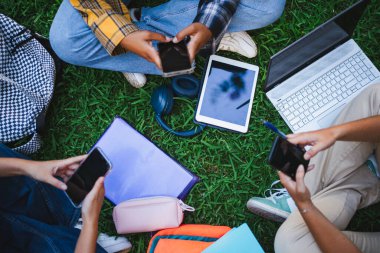 Üniversite öğrencilerinin otların üzerinde otururken ellerini telefonlarına koydukları yüksek açılı bir fotoğraf.