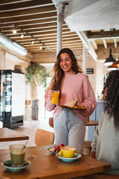 Brunch restoranındaki masaya yemeğini taşıyan gülümseyen genç bir kadın..