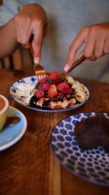 Kahve dükkanında çilekli, muzlu ve yaban mersinli waffle kesen bir el resmi..