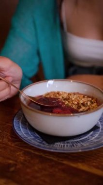 Genç bir kadının bir kaşık dolusu ahududu ve tahıl gevreğiyle acai kasesini kepçelemesi.