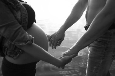 Hamile bir kadın ve partnerinin, suyun kenarında nazikçe el ele tutuşup sevgi ve beklentiyle dolu samimi ve duygusal bir anı yakaladıkları siyah beyaz bir fotoğraf..