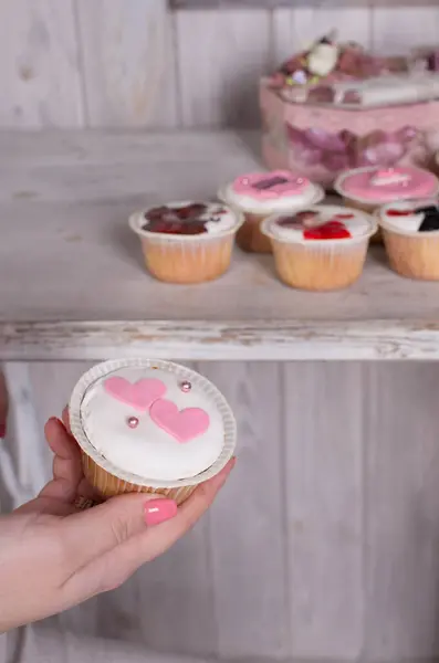 Süslü bir kek, beyaz krema, pembe fontane kalpler ve şeker incileri olan bir el resmi. Yaratıcı tasarımları olan diğer kapkekler arka planda bulanıklaşıyor. Tatlı, romantik ve neşeli tatlı konsepti.