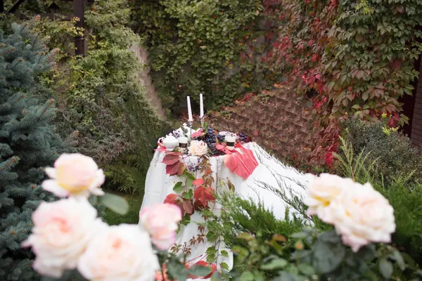 Mumlar, üzümler, çiçekler ve çay fincanlarıyla süslenmiş küçük bir açık hava masası. Etrafı yeşillik ve güllerle çevrili, romantik ve rahat bir bahçe atmosferi yaratıyor..