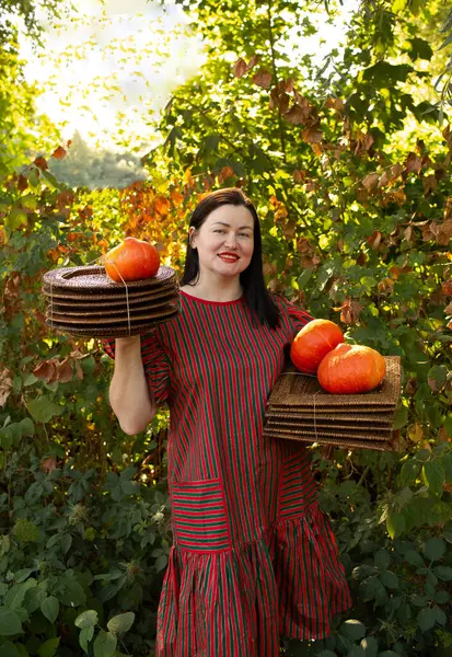 Elinde balkabağı ve doğal hasır tepsiler tutan çizgili eko tarzı bir elbise giymiş bir kadının portresi. Sürdürülebilir ürünlerle kırsal sonbahar kavramı.