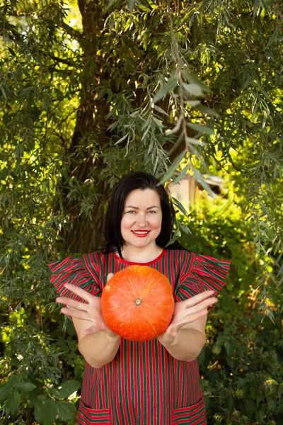 Bir ağacın altında elinde parlak turuncu bir balkabağı tutan bir kadın. Sonbahar hasadı ve sürdürülebilirlik teması, eko yaşamı, doğal tarım ve sağlıklı organik gıdanın sembolü.