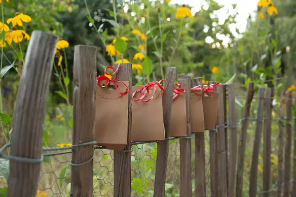 Etrafı yeşilliklerle çevrili kırmızı kurdeleli bir bahçe çitine bağlanmış bir sürü kraft kağıdı hediye paketi. Doğal ve ekolojik ambalaj fikri.